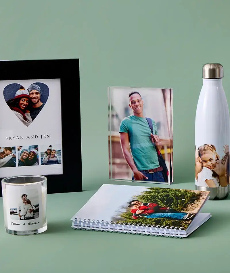 A display of personalised photo gifts including framed prints, a photo candle, a spiral photo notebook, a photo water bottle and a photo block arranged on a green surface.